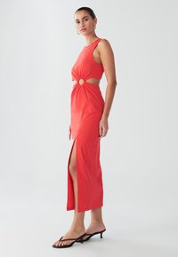 Crvena haljina bez rukava s izrezom u struku, naglašena okruglim detaljem. Ima visoki prorez na bedrima i uski kroj.