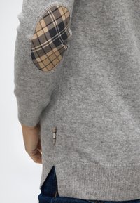 Manche de pull gris avec un patch de coudes à carreaux beige et noir sur une personne portant un jean bleu foncé.