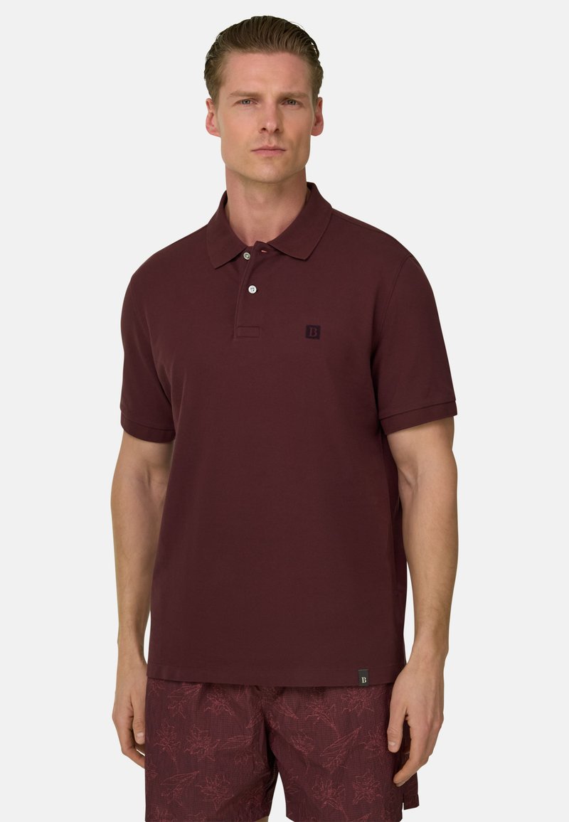 Homme portant un polo à manches courtes marron rouge et un short à motifs floraux marron rouge, debout devant un fond clair uni.