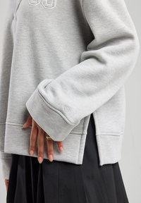 Harmaa huppari, jossa on ribattu hihansuu ja sivureikä, ja jossa on brodeerattu logo rinnassa. Sujuva rakenne, rento muotoilu.