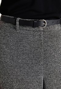 Pantalons texturés gris et noirs avec une ceinture en cuir noir. La ceinture est dotée d'une boucle ronde en argent qui complète le motif du tissu.