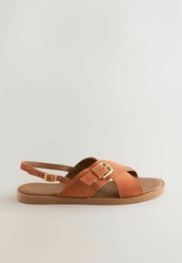 Sandale en daim couleur cognac avec des brides croisées et une boucle réglable, dotée d'une semelle plate et d'une doublure intérieure lisse de couleur marron clair. Design minimaliste.