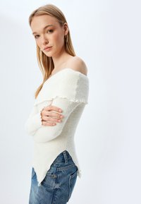 Maglione bianco scollato sulle spalle con lavorazione a maglia testurizzata, bordo arricciato e orlo asimmetrico, abbinato a jeans blu.