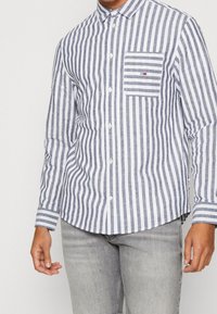 Koszula w paski w kolorze niebiesko-białym, z kieszenią na piersi, długimi rękawami i zaokrąglonym dołem. Wykonana z lekkiego materiału.