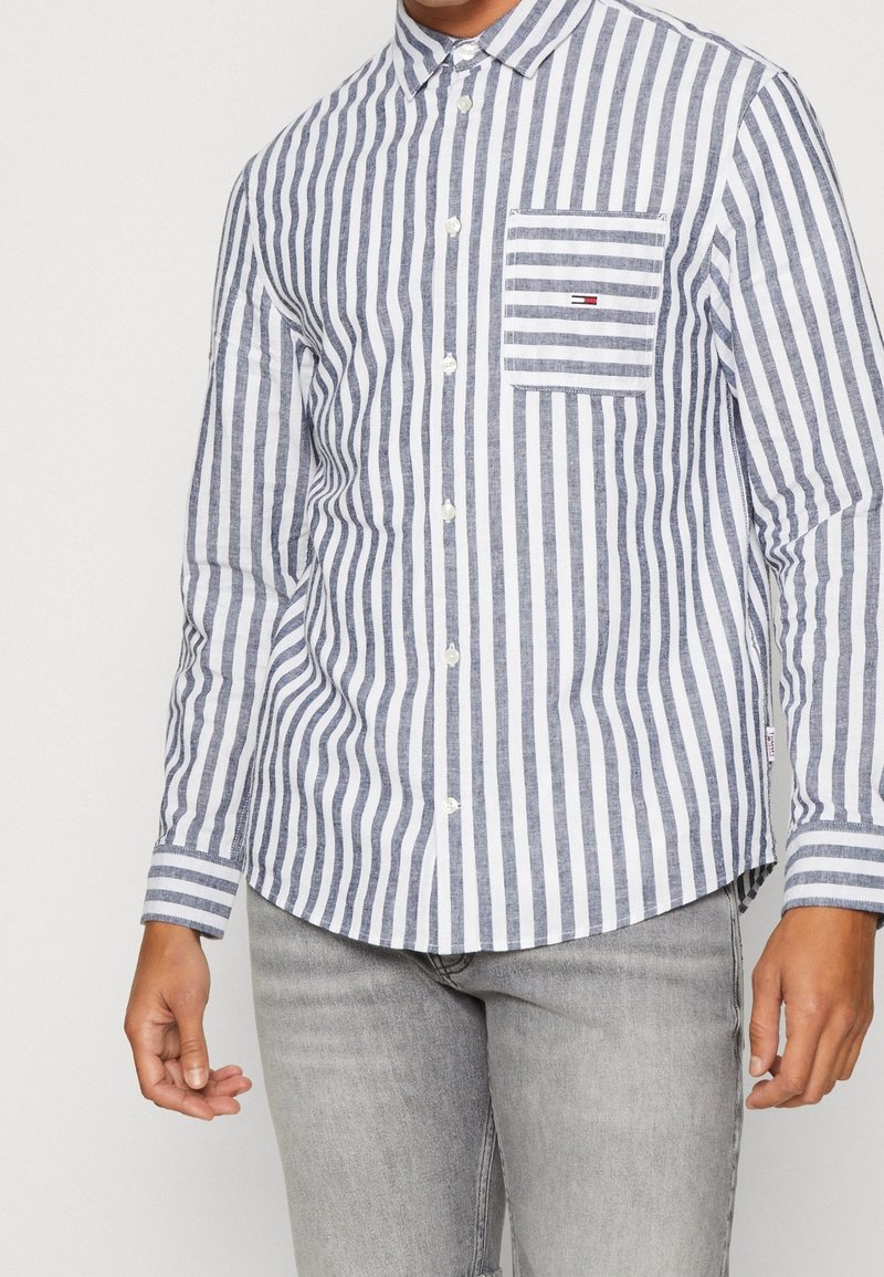 Koszula w paski w kolorze niebiesko-białym, z kieszenią na piersi, długimi rękawami i zaokrąglonym dołem. Wykonana z lekkiego materiału.