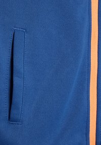 Blauer Sweatshirtstoff mit einer glatten Textur, versehen mit einem orangen vertikalen Streifen und einer Seitentasche. Einfaches Design mit klaren Linien.
