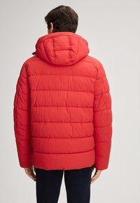 Giacca a vento rossa con cappuccio, caratterizzata da una texture trapuntata e polsini elasticizzati. Il materiale appare liscio e leggero, progettato per il calore.