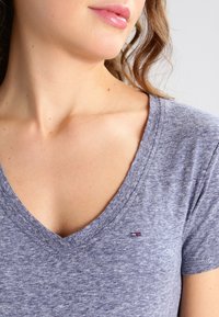 Grå v-ringad t-shirt i mjukt, strukturerat tyg. Har en liten, märkeslogotyp på vänster bröst. Närbild av halsringning och axel.