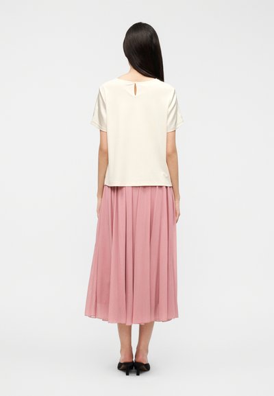 Μπλούζα κοντομάνικη, κρεμ, με λεπτομέρεια κλειδαρότρυπας στην πλάτη, συνδυασμένη με ροζ πλισέ μίντι φούστα. Το ύφασμα φαίνεται ελαφρύ και αέρινο.