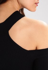 Frau in einem schwarzen Rippoberteil mit ausgeschnittenen Schultern und asymmetrischem Dekolleté, das Haut zeigt und eine teilweise Seitenansicht zeigt.