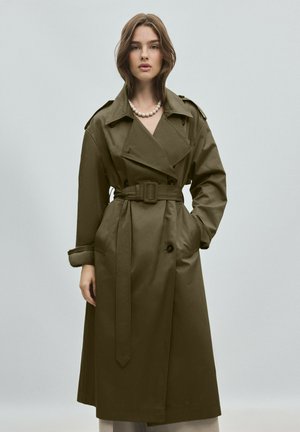 Jeune femme portant un long trench-coat vert olive ceinturé et un collier de perles, debout devant un fond uni gris clair.