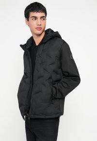 Sort puffjakke med glatt finish, hette, glidelåslukking og elastiske mansjetter. Har kontrasterende matte sider og sidelommer.