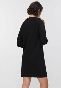 Robe pull noire à coupe décontractée, manches longues, design à épaules dénudées et détails décoratifs à clous au niveau du col. Texture lisse.