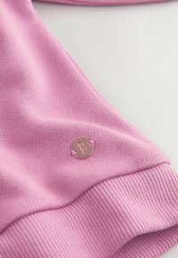Sudadera rosa claro con un tejido texturizado, puños acanalados y un pequeño logotipo metálico cerca del dobladillo.