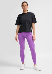 Svartprickig kortärmad topp i kombination med tajta lila leggings och vita träningsskor, stående mot en neutral bakgrund.