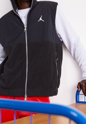 Personne portant un gilet noir à fermeture éclair avec le logo Jumpman blanc par-dessus une chemise à manches longues blanche, debout à côté d'une table en bois et d'une chaise bleue.