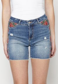 Denim-shortsit, joiden helmat ovat revityt ja joissa on punaiset kukka-ompelukset sivusaumoissa, yhdistettyinä tiukkaan valkoiseen hihattomaan toppiin.