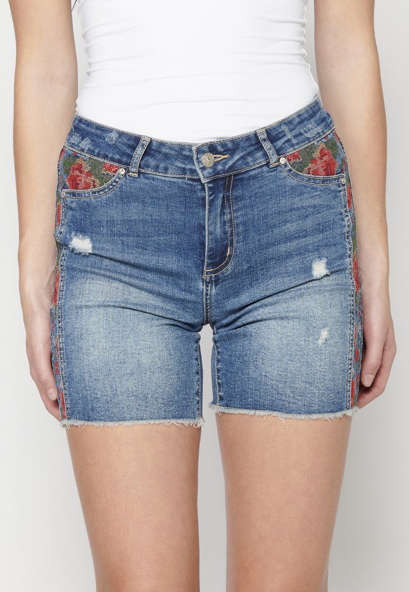 Denim-shortsit, joiden helmat ovat revityt ja joissa on punaiset kukka-ompelukset sivusaumoissa, yhdistettyinä tiukkaan valkoiseen hihattomaan toppiin.