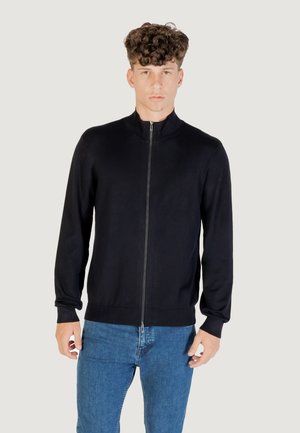 Giovane con capelli ricci che indossa un maglione nero con zip e jeans blu, su uno sfondo chiaro e semplice.