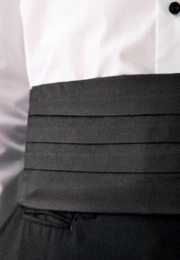 Camisa blanca de vestir metida dentro de pantalones de esmoquin negros con un fajín negro texturizado alrededor de la cintura y un botón negro visible.