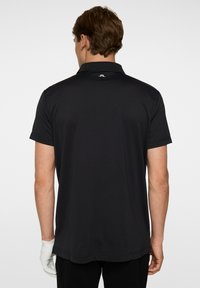 Musta polo-paita lyhyillä hihoilla, sileä pinta ja hillitty kaulus. Ylhäällä selässä pieni logo.