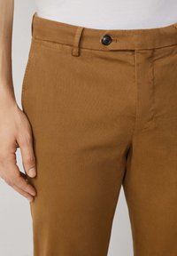 Pantalones de algodón marrón con una textura suave, que cuentan con un cierre de botón, trabillas para cinturón y un corte ajustado. Detalles de costura visibles en los laterales.
