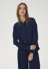 Camicia bottonata blu navy con colletto alla cinese, maniche lunghe e vestibilità rilassata. Tessuto liscio e dettagli drappeggiati sottili.