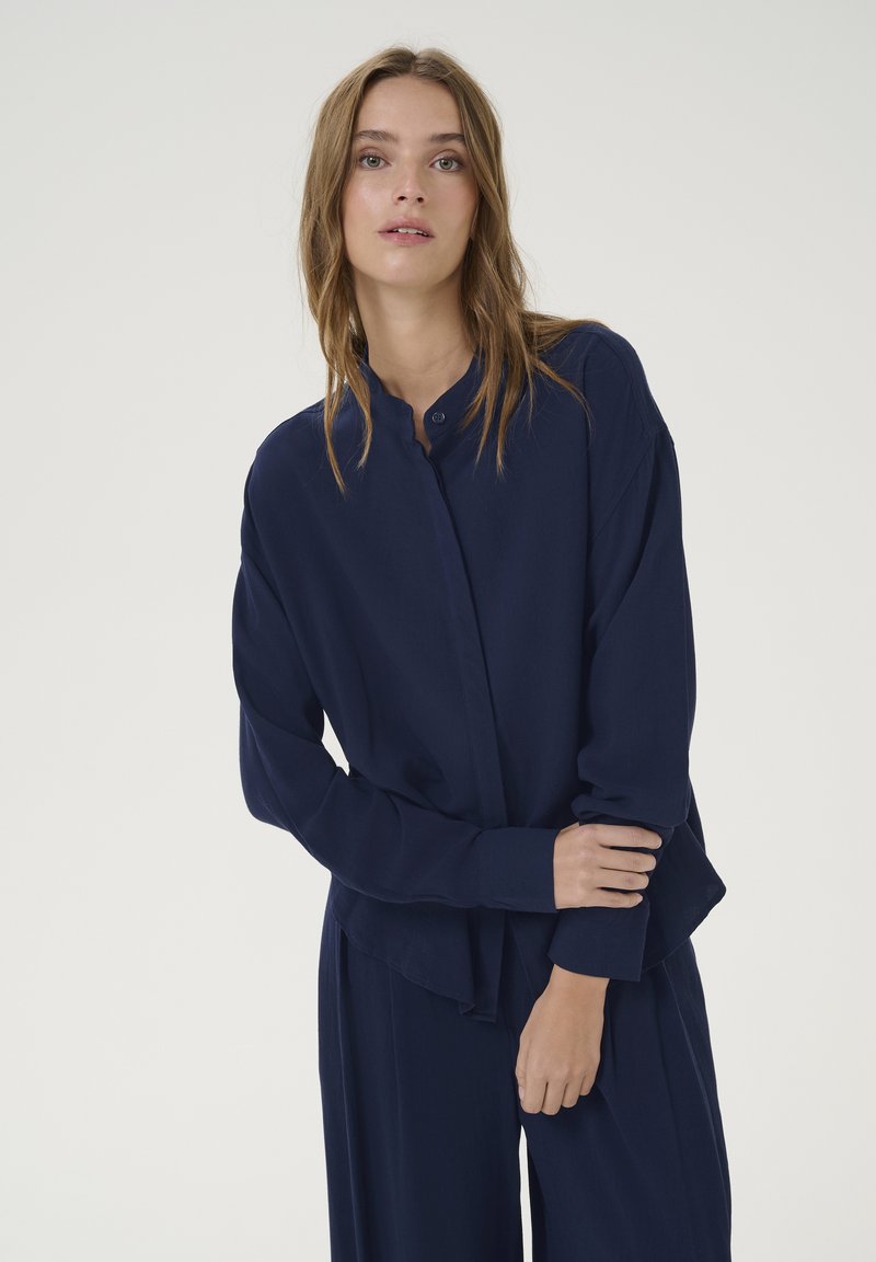 Camicia bottonata blu navy con colletto alla cinese, maniche lunghe e vestibilità rilassata. Tessuto liscio e dettagli drappeggiati sottili.