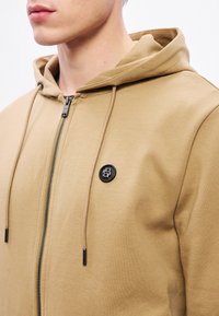 Béžová mikina na zips s klokankovým vreckom, šnúrkami a čiernou nášivkou s logom. Materiál pôsobí mäkko s hladkým povrchom.