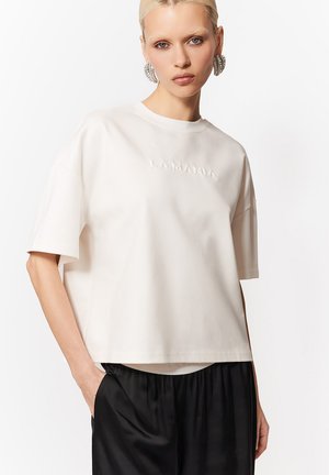 Kobieta o gładko zaczesanych blond włosach, ubrana w biały top z krótkim rękawem z haftowanym logo i czarne spodnie, stoi na tle białego tła.