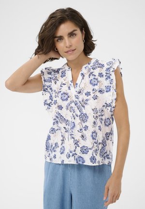 Kvinde med kort brunt hår iført en hvid ærmeløs bluse med blåt blomsterprint og lyseblå bukser, poserer med den ene hånd, der rører ved håret.