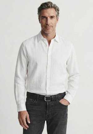 Homme aux cheveux gris portant une chemise blanche boutonnée rentrée dans un jean noir avec une ceinture noire tressée, une main dans la poche, fond neutre.