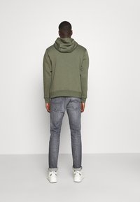 Olivgrön hoodie med huva, figursydd vid midjan, kombinerad med grå jeans. Vita sneakers med detaljer. Enkelt bakgrund, neutralt färgschema.