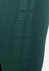 Leggings verde scuro con un motivo testurizzato sottile e il marchio ripetuto lungo il lato. Realizzati in materiale elastico e liscio.