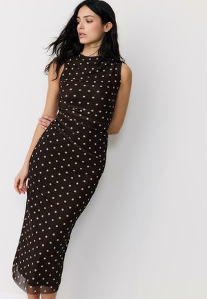 Ženska z dolgimi temnimi lasmi, oblečena v črno midi obleko brez rokavov z bež pikami, stoji ob belem zidu in gleda v stran.