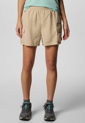 Donna che indossa pantaloncini cargo beige con vita elastica, scarpe da trekking grigie e maglietta verde menta, in piedi su sfondo grigio.
