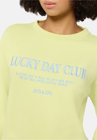 Jasnożółta bluza z haftowanym niebieskim napisem: "LUCKY DAY CLUB" oraz cytatem poniżej. Gładki materiał, luźny krój i okrągły dekolt.
