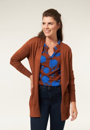 Vrouw met een bruine cardigan over een blauw bloesje met bloemen en een donkere spijkerbroek, glimlachend en naar de zijkant kijkend met één hand op haar heup.