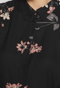 Mustat pusero, jossa on vaaleanpunaisia ja harmaita kukkakuvioita, terävä kaulus ja mustista kankaista päällystettyjä nappeja edessä.