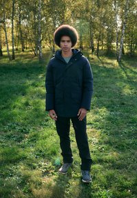 Navy puffer jacket with a hood, two front pockets, worn over a gray shirt, paired with black pants and gray hiking shoes on grass.
