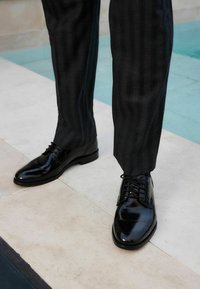 Chaussures habillées en cuir noir avec une finition brillante et un laçage, assorties à un pantalon à rayures fines de couleur sombre, posées sur un sol en carrelage clair.