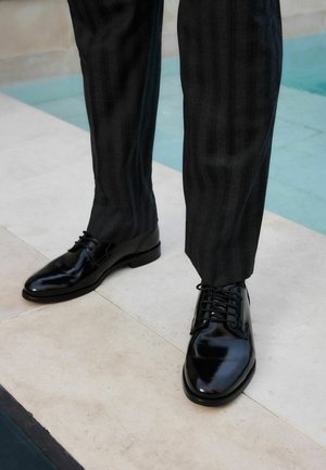 Zapatos de vestir de cuero negro con un acabado brillante y diseño con cordones, combinados con pantalones oscuros a rayas, exhibidos sobre una superficie de baldosas claras.