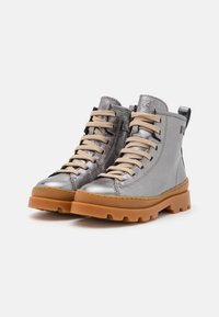 Botas de tobillo de cuero plateado con cordones beige, superficie texturizada y suela de goma con un patrón de agarre robusto. Presenta una lengüeta en el talón.