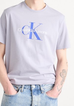 Osoba ubrana w jasnofioletowy t-shirt Calvin Klein Jeans z niebieskim logo oraz jasnoniebieskie dżinsy, z rękami w kieszeniach, widoczne tatuaże na ramionach.