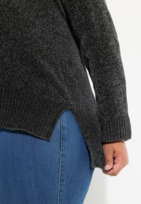 Donkergrijze gestructureerde gebreide trui met geribde zoom, zijsplitten en lange mouwen, gelaagd over blauwe denim met zichtbare stiksels.