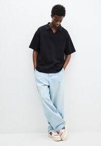 Polo shirt nero a maniche corte con vestibilità rilassata, abbinato a jeans azzurri larghi e sneakers bianche con dettagli rossi e neri.