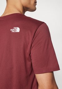Burgunderfarget t-skjorte med korte ermer laget av mykt stoff, med en hvit logo på øvre del av ryggen og en klassisk rund hals.