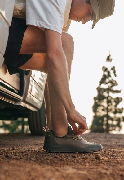 Zapatillas deportivas grises con parte superior de malla y acentos negros. Suela de goma, tira en el talón. Hombre atándose los cordones mientras está sentado en un vehículo.