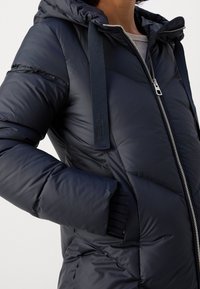 Chaqueta acolchada de color azul marino con capucha, que presenta un cierre frontal con cremallera, correas en contraste y una textura acolchada. Incluye bolsillos laterales y un acabado brillante.