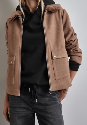 Person iført en tan lynlåslukket uldjakke med klaplommer over en sort hættetrøje og mørke jeans mod en ens grå baggrund.